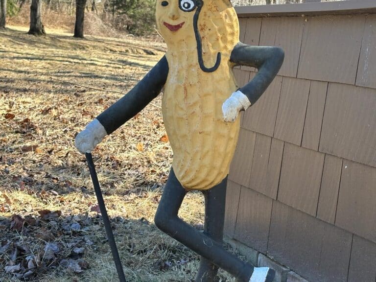 The Cast Iron "Life Size" Mr. Peanut Statue, featuring a monocle and cane, is standing on one leg outside near a small building.