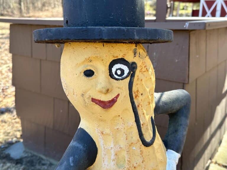 The Cast Iron "Life Size" Mr. Peanut Statue stands outside a wooden structure, featuring a peanut character in a top hat and monocle with one arm akimbo.