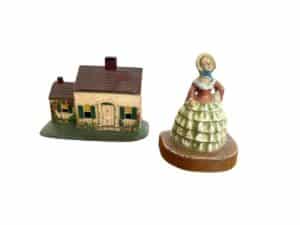 A cast iron painted Southern lady in a dress stands beside a Cape Cod cottage model door stop, set against a white background.