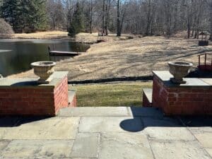 Stone steps lead to a pond with a wooden dock, surrounded by bare trees and grass. A pair of Stone Garden Urns Planters sit on the steps.