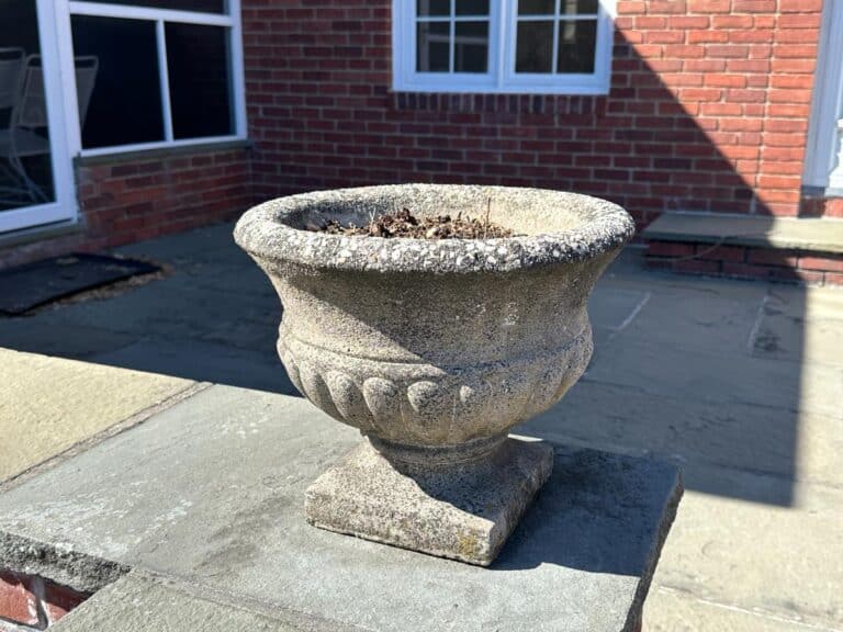 Stone planters with soil, placed on a patio in front of a brick house with windows, from the Pair of Stone Garden Urns Planters collection.