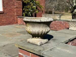 A Pair of Stone Garden Urns Planters sits on a brick patio, flanked by bare trees and a red brick building.