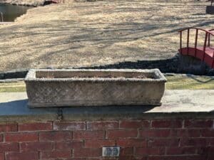 Vintage concrete planters on a brick ledge bask in sunlight, with a grassy field and red bridge in the backdrop.
