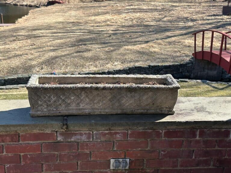 Vintage concrete planters on a brick ledge bask in sunlight, with a grassy field and red bridge in the backdrop.