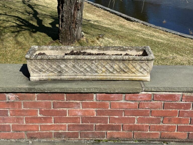 Set atop a brick wall near grass and water, the Vintage Rectangular Concrete Planters come filled with soil.