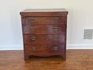 A Bachelor's Chest of Drawers, featuring four brass-handled drawers, stands against a white wall on a hardwood floor.