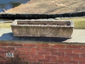 Vintage rectangular concrete planter from a set of three, filled with soil and positioned on a brick wall near grass and a pond.