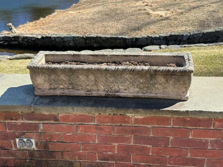 Vintage rectangular concrete planter from a set of three, filled with soil and positioned on a brick wall near grass and a pond.