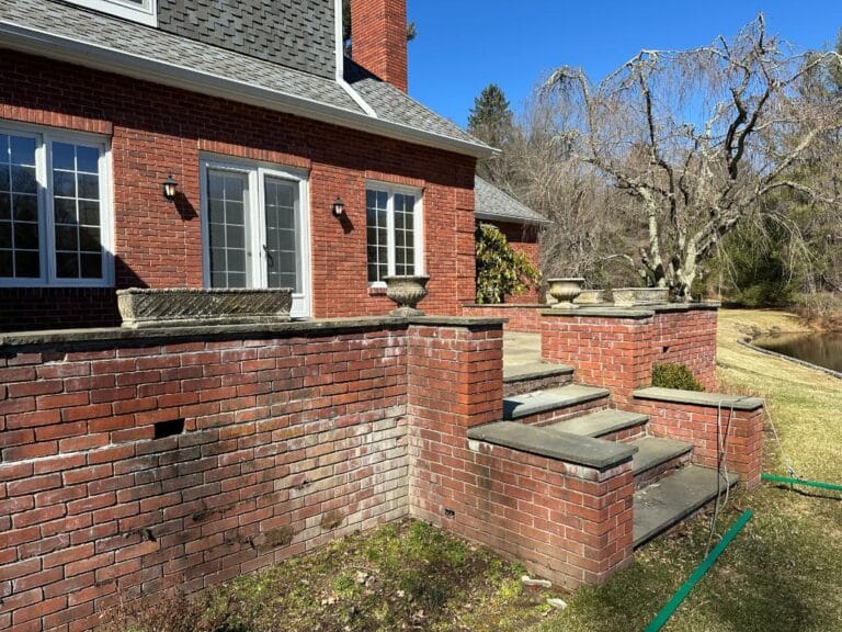 Brick house with steps leading to a raised patio, adorned with a set of three vintage rectangular concrete planters, surrounded by grass and trees under a clear blue sky.