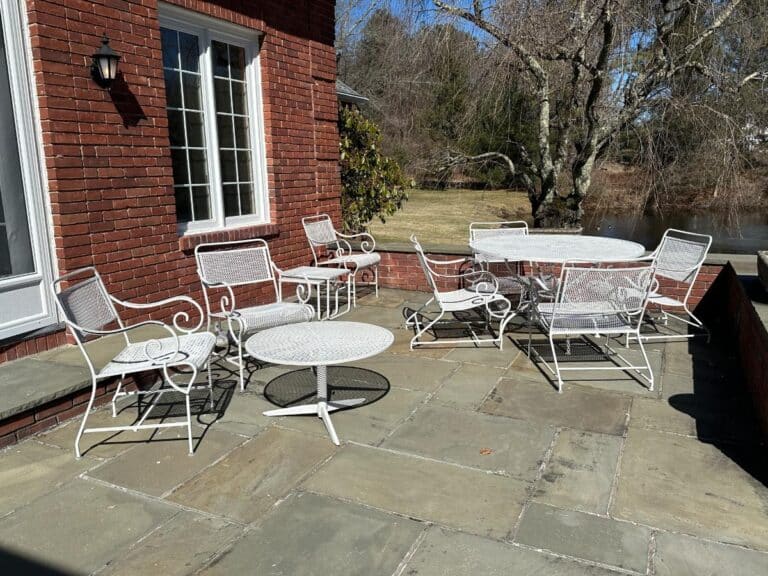 The Vintage White Wrought Iron Outdoor Dining and Conversation Set is placed on a stone terrace surrounded by brick walls, with bare trees in the background.