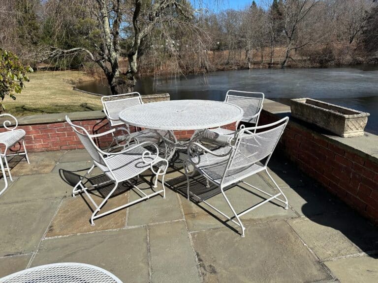 Vintage White Wrought Iron Outdoor Dining and Conversation Set on a stone terrace by a pond with bare trees in the background.