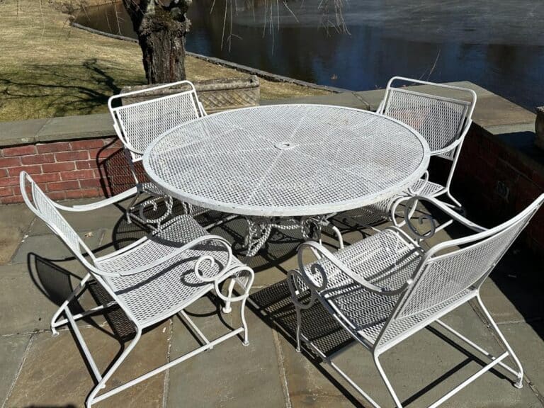 A Vintage White Wrought Iron Outdoor Dining and Conversation Set on a stone terrace by the water, surrounded by bare trees and grass.