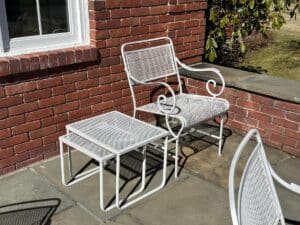 The Vintage White Wrought Iron Outdoor Dining and Conversation Set sits on a sunlit brick patio beside a grassy lawn.