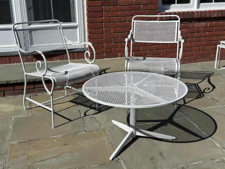 A Vintage White Wrought Iron Outdoor Dining and Conversation Set featuring a round table and two metal chairs is placed on a stone patio against a brick wall.