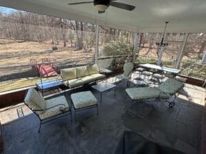Enjoy a screened patio featuring a Vintage Wrought Iron Patio Set with newly upholstered cushions and tables under a ceiling fan, overlooking a yard with a red bridge crossing over a stream.