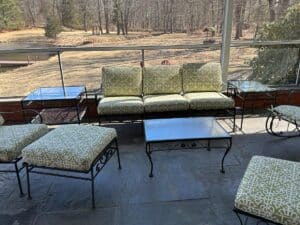 A vintage wrought iron patio set with newly upholstered patterned green cushions overlooks a dry garden and pond.