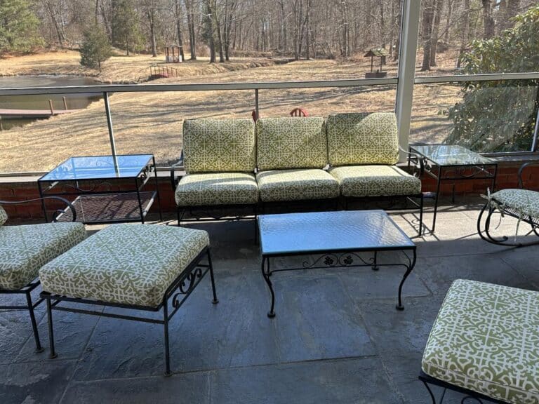 A vintage wrought iron patio set with newly upholstered patterned green cushions overlooks a dry garden and pond.