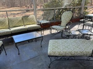 A Vintage Wrought Iron Patio Set featuring newly upholstered green patterned cushions rests on a stone floor, offering views of a wooded area through glass panels.