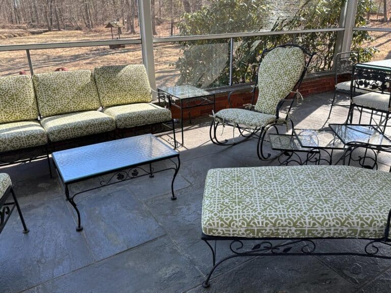 A Vintage Wrought Iron Patio Set featuring newly upholstered green patterned cushions rests on a stone floor, offering views of a wooded area through glass panels.