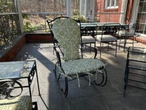 A patio with a vintage wrought iron set, including a rocking chair and table with newly upholstered green patterned cushions, is bathed in sunlight.