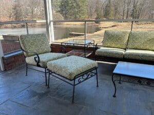 The Vintage Wrought Iron Patio Set, featuring newly upholstered cushions and elegant tables, overlooks a pond with trees in the background.