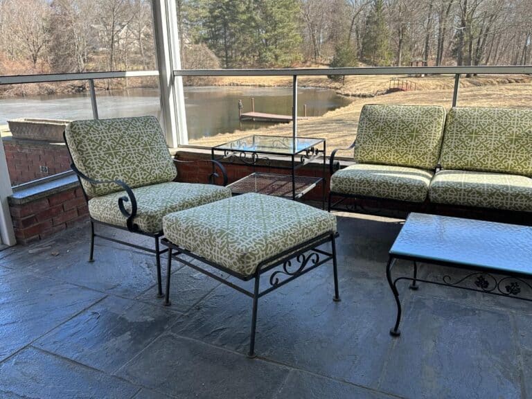The Vintage Wrought Iron Patio Set, featuring newly upholstered cushions and elegant tables, overlooks a pond with trees in the background.