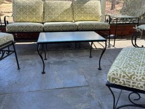 Vintage Wrought Iron Patio Set with newly upholstered green patterned cushions and a metal table on a stone floor, against a red brick wall.