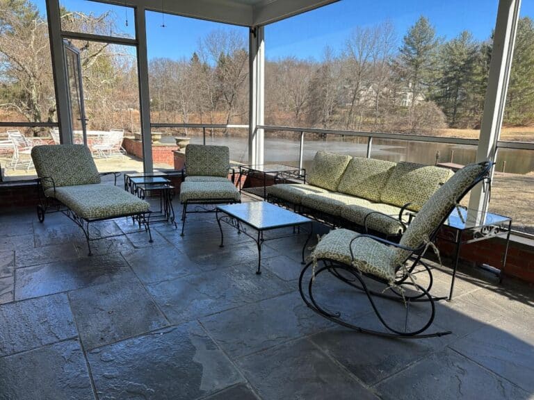 A sunlit patio features a Vintage Wrought Iron Patio Set with newly upholstered green cushions, complemented by glass tables, overlooking a pond and bare trees.