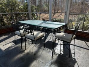 The Vintage Wrought Iron Grapevine Dining Set, featuring a glass-top table and six chairs with seat cushions, is perfect for a sunlit patio surrounded by greenery and trees.