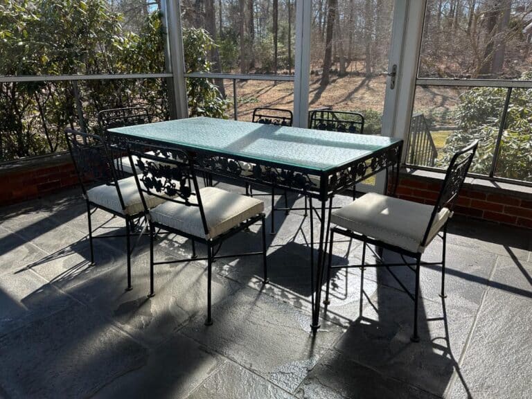 The Vintage Wrought Iron Grapevine Dining Set, featuring a glass-top table and six chairs with seat cushions, is perfect for a sunlit patio surrounded by greenery and trees.