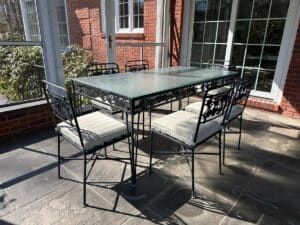 A Vintage Wrought Iron Grapevine Dining Set with seat cushions, featuring a glass-top table and six metal chairs, graces the sunlit patio surrounded by brick walls and windows.