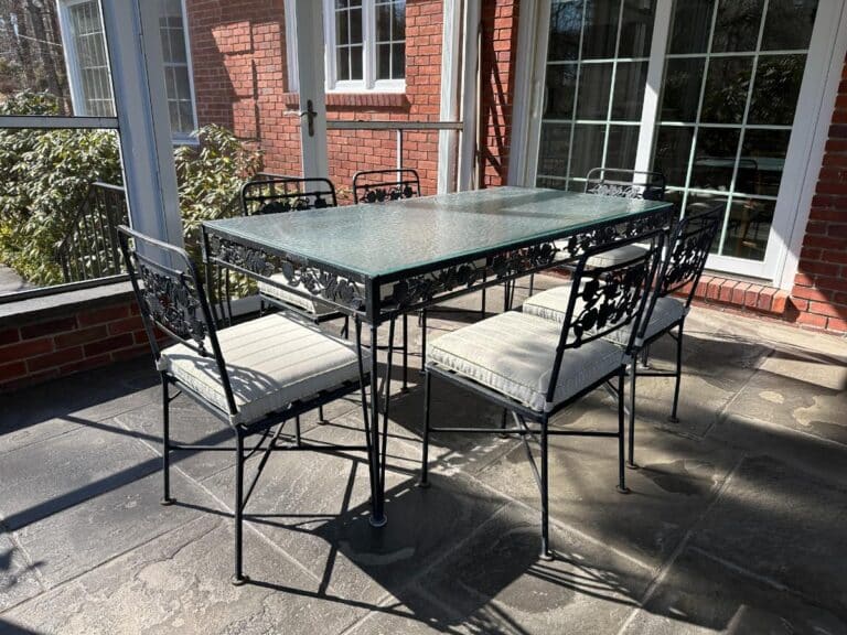 A Vintage Wrought Iron Grapevine Dining Set with seat cushions, featuring a glass-top table and six metal chairs, graces the sunlit patio surrounded by brick walls and windows.