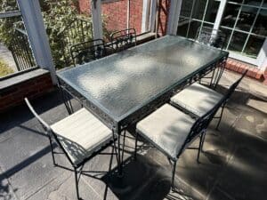 The sunlit stone floor features a Vintage Wrought Iron Grapevine Dining Set with six chairs and seat cushions, situated near a red brick wall and window.