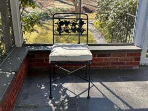 The Vintage Wrought Iron Grapevine Dining Set with Seat Cushions, featuring a floral backrest, sits on a sunlit porch with a brick wall and garden view.