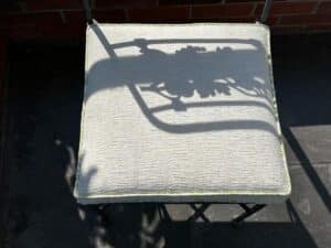 A metal chair from the Vintage Wrought Iron Grapevine Dining Set casts a shadow with a leaf design on a sunlit, light-colored cushion.