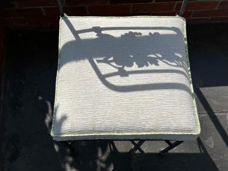 A metal chair from the Vintage Wrought Iron Grapevine Dining Set casts a shadow with a leaf design on a sunlit, light-colored cushion.