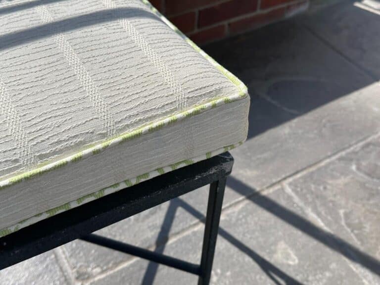 Close-up of a textured white cushion with green trim from the Vintage Wrought Iron Grapevine Dining Set, on a black metal frame, against a stone floor and brick wall.