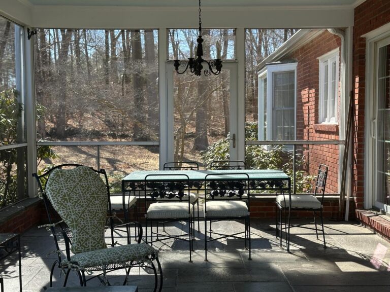 Screened porch furnished with the Vintage Wrought Iron Grapevine Dining Set, a chandelier, and a cushioned lounge chair as sunlight filters through bare trees.