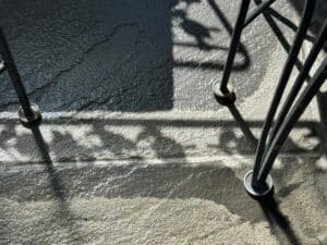 Sunlight and shadows play on the wet, textured concrete floor beneath the Vintage Wrought Iron Grapevine Dining Set with seat cushions.