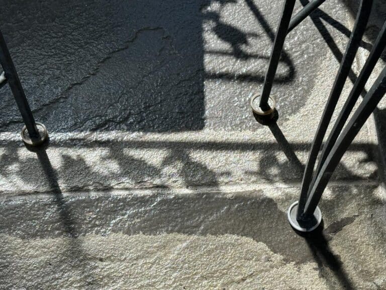 Sunlight and shadows play on the wet, textured concrete floor beneath the Vintage Wrought Iron Grapevine Dining Set with seat cushions.