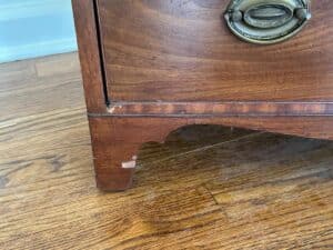 Close-up of a Bachelor's Chest of Drawers corner with a brass handle, displaying slight wear and scuff marks on a wooden floor.