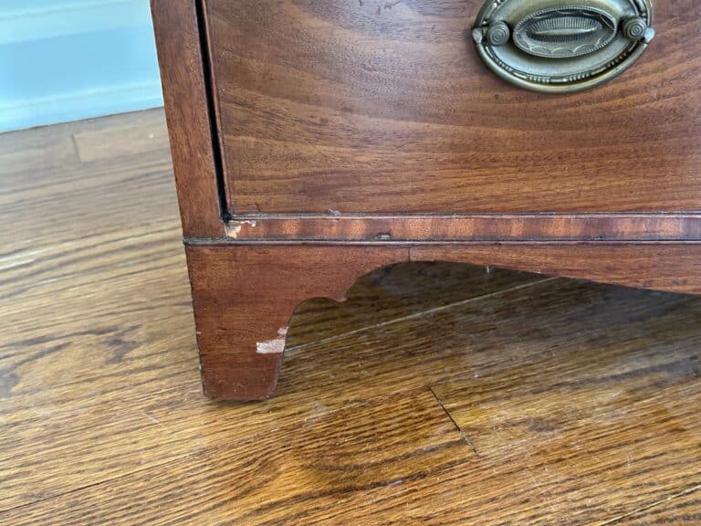 Close-up of a Bachelor's Chest of Drawers corner with a brass handle, displaying slight wear and scuff marks on a wooden floor.