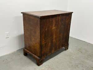 An Antique Burl Chest of Drawers with a scratched surface is positioned in a corner of a room featuring pale walls and a concrete floor.