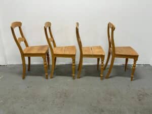 Four wooden chairs with rounded backs are placed in a line on a concrete floor against a white wall.