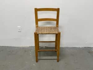 A wooden stool with a woven seat, set against a plain white wall and gray concrete floor.