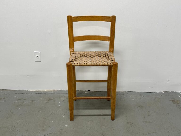 A wooden stool with a woven seat, set against a plain white wall and gray concrete floor.