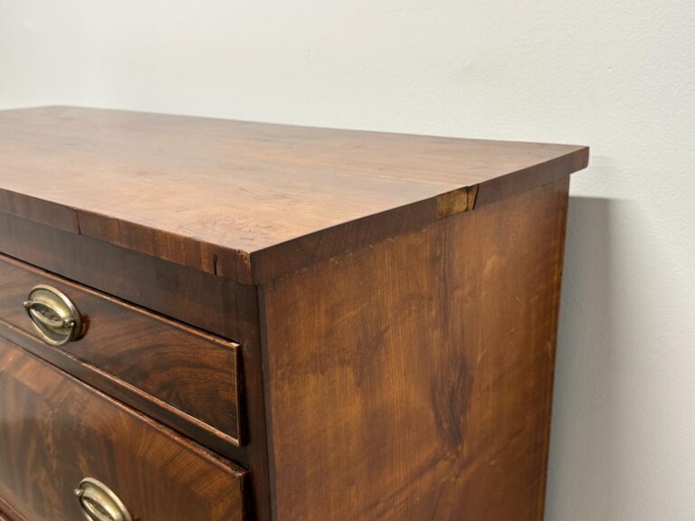 Close-up of a wooden dresser corner with drawers and brass handles against a light-colored wall.