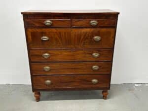 A vintage wooden chest of drawers with five drawers and metal handles, standing on short legs against a white wall.