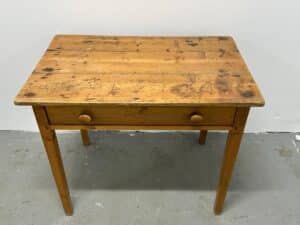 A small, weathered wooden table with a single drawer and two knobs against a plain white wall.