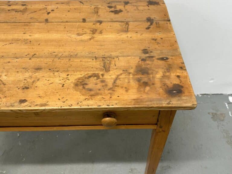 Wooden table with visible stains and a small drawer, set against a gray floor and white wall.